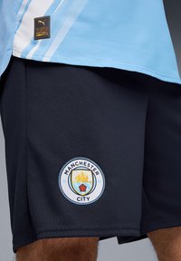 Shorts Manchester City en tissu bleu marine foncé, avec un logo brodé rond comportant un bateau et une rose, surface texturée et taille élastique.