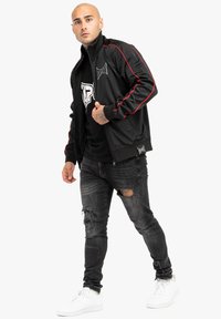 Chaqueta atlética negra con acentos rojos, cierre de cremallera y logo; combinada con una camiseta gráfica negra y jeans negros desgastados. Zapatillas blancas.