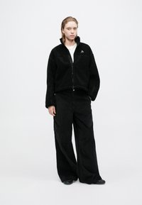 Jachetă din fleece neagră, cu fermoar și guler înalt, combinată cu pantaloni largi din catifea cord neagră, dotate cu buzunare laterale. Design simplu, material texturat.