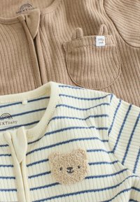 Dos bodies de bebé de canalé, uno beige con un bolsillo con orejas pequeñas y etiqueta "little cub", el otro crema con rayas azules y un parche con cara de osito.