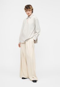 Άτομο που φοράει μια φαρδιά μπεζ πουκάμισο με γιακά και εκρού παντελόνια με φαρδιά πόδια, στέκεται με το ένα χέρι στην τσέπη σε απλό φόντο.