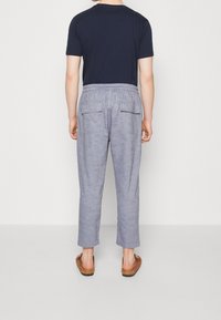 Šedé lněné kalhoty se stahovací šňůrkou v pase, dvěma zadními kapsami a manžetami. Nošeny s tmavě modrým tričkem a sandály.