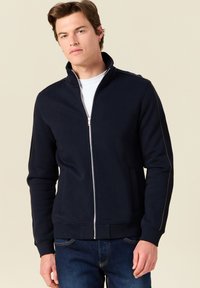 Giacca a zip blu navy realizzata in tessuto morbido con colletto alto, caratterizzata da leggeri dettagli di cucitura lungo le spalle e due tasche frontali.