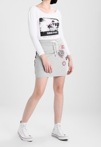 Langärmeliges weißes Hemd mit grafischem Aufdruck, kombiniert mit einem grauen Minirock mit bunten Aufnähern und Kordelzugbund, getragen mit weißen Turnschuhen.