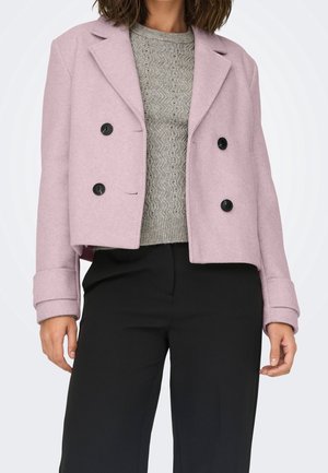 Femme portant un manteau court croisé rose, un pull en maille grise texturée et un pantalon noir taille haute sur un fond uni.