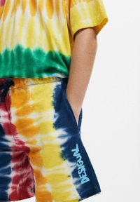 Maglietta tie-dye in tonalità di giallo, verde, arancio e rosso; abbinata a pantaloni corti blu con una tasca e il marchio "Desigual" in azzurro chiaro.