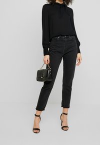 Blusa nera con dettaglio a fiocco, jeans neri a vita alta, tacchi neri con cinturino e una borsa nera strutturata con tracolla a catena.