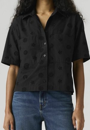 Kvinde iført en sort kortærmet skjorte med knapper og broderede blomsterdesigns, parret med blå denimjeans.