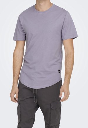 Mand iført en lyse lilla kortærmet T-shirt med rund hals og mørkegrå cargobukser med lommer, stående mod en ensfarvet baggrund.