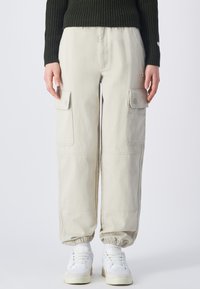 Beige Cargo-Hosen mit elastischen Bündchen, ausgestattet mit mehreren Seitentaschen und einem lässigen Schnitt, kombiniert mit weißen Sneakers.
