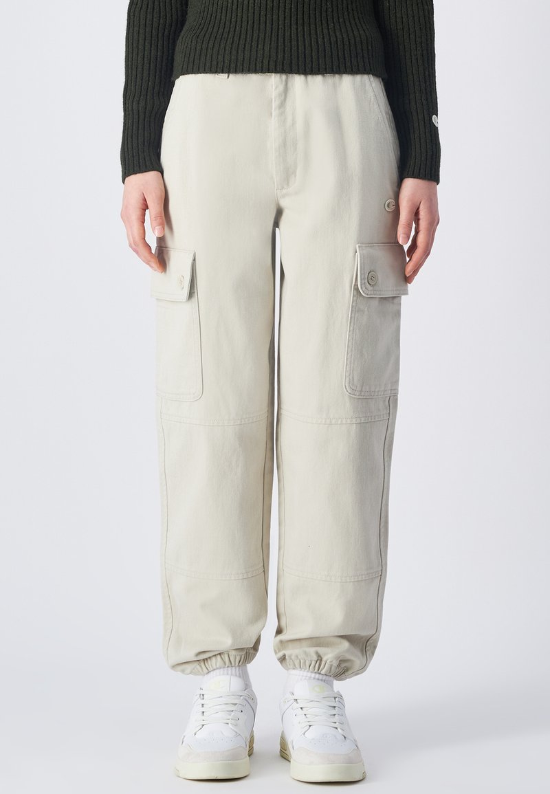 Beige Cargo-Hosen mit elastischen Bündchen, ausgestattet mit mehreren Seitentaschen und einem lässigen Schnitt, kombiniert mit weißen Sneakers.