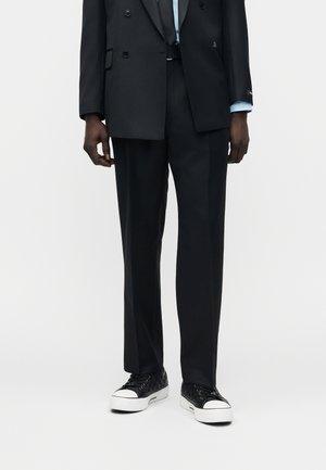 Mees, kes kannab musti pükse, musta topeltrinnalist bleiserit, helerohelisi särgivarrukaid ning musti tossusid valgete talladega.