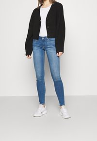 Cárdigan negro de punto acanalado con botones, combinado con jeans ajustados azules y zapatillas blancas. El conjunto incluye una simple camiseta blanca debajo.