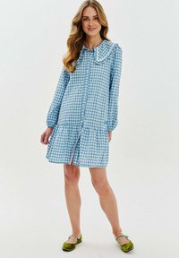 Svetlomodré kockované šaty s volánovým golierom, predným zapínaním na gombíky, dlhými rukávmi a sukňou v vrstevnatom strihu. Nosené s zelenými balerínkami.