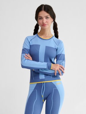 Młoda kobieta z warkoczami, ubrana w niebiesko-żółtą bluzkę z długim rękawem i dopasowane legginsy, stoi ze skrzyżowanymi ramionami na białym tle.