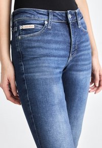 Vaqueros de mezclilla azul con un diseño ajustado, un ligero desgaste, cinco bolsillos y una etiqueta de Calvin Klein en el bolsillo delantero.