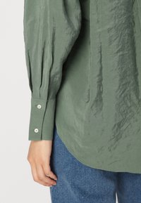 Langermet skjorte i salviegrønn med teksturert stoff, knappelinning ved ermene og buet kant, sammen med lyseblå denim.