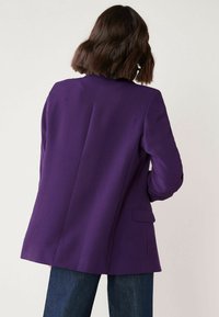 Blazer violet avec une coupe ajustée, des revers crantés et une fente unique au dos. Confectionné dans un tissu lisse avec une silhouette structurée.