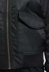 Schwarze Bomberjacke mit einem goldenen Reißverschluss, einer Druckknopf-Tasche an der Seite und gerippten Bündchen für zusätzliche Textur.