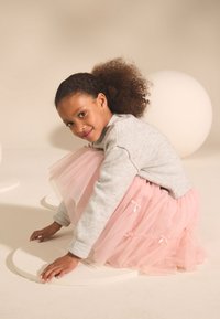 Sivý pletený sveter, ružová tylová sukňa s mašľami, dieťa skrčené na kruhovom predmete, svetlé pozadie, mäkké textúry a jemné farby.