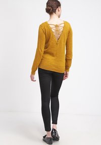 Mosterdgele trui met een diepe V-achterkant en vetersluiting, gecombineerd met zwarte leggings en platte zwarte schoenen. Soepele textuur.