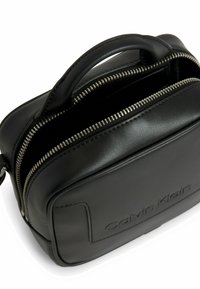 Černá kožená kabelka se strukturovaným tvarem, horním uchem a stříbrným zipem. Na předním panelu má vyražené logo značky.