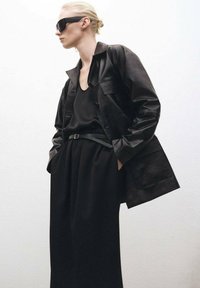 Black leather jacket with a relaxed fit, featuring buttons, a chest pocket, and a smooth finish, paired with a loose black top and wide trousers.