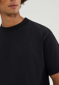 Schwarzes T-Shirt aus Baumwolle mit Rundhalsausschnitt, kurzen Ärmeln und einem kleinen geprägten Logo auf der Brust. Glatte Textur.