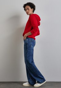 Felpa rossa con una vestibilità rilassata, abbinata a jeans blu a gamba larga e sneakers bianche. L'outfit è casual e confortevole, con accenti minimali.