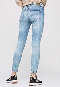 Personne vêtue d'un jean skinny bleu clair, d'un pull rayé beige et de baskets noir et blanc, se tenant devant un fond uni.