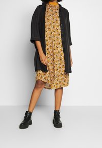 Černá oversized košile s knoflíky, překrytá přes žluté květinové šaty, doplněná černými kotníkovými botami se sponkovými detaily.