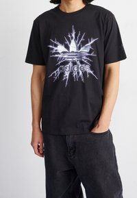 Černé bavlněné tričko s velkým logem Adidas a elektrickými bleskovými akcenty v bílé a šedé barvě. Volný střih, krátké rukávy, casual design.
