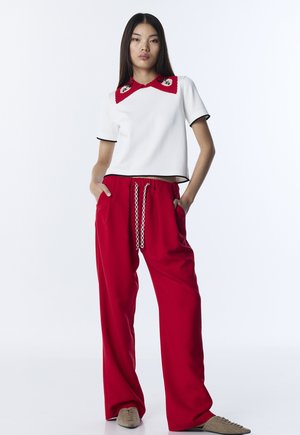 Młoda kobieta w białym krótkim rękawie z czerwonym kołnierzem i szerokich czerwonych spodniach na sznurku, stojąca z rękami w kieszeniach na białym tle.