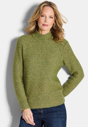 Pull en tricot vert avec un col montant, tissu texturé, poignets en côtes et ceinture en côtes. Porté avec un jeans bleu foncé.