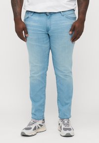Jeans de mezclilla de color azul claro con un ajuste slim, con cremallera clásica, cinco bolsillos y una sutil textura deslavada. Combinados con zapatillas grises.