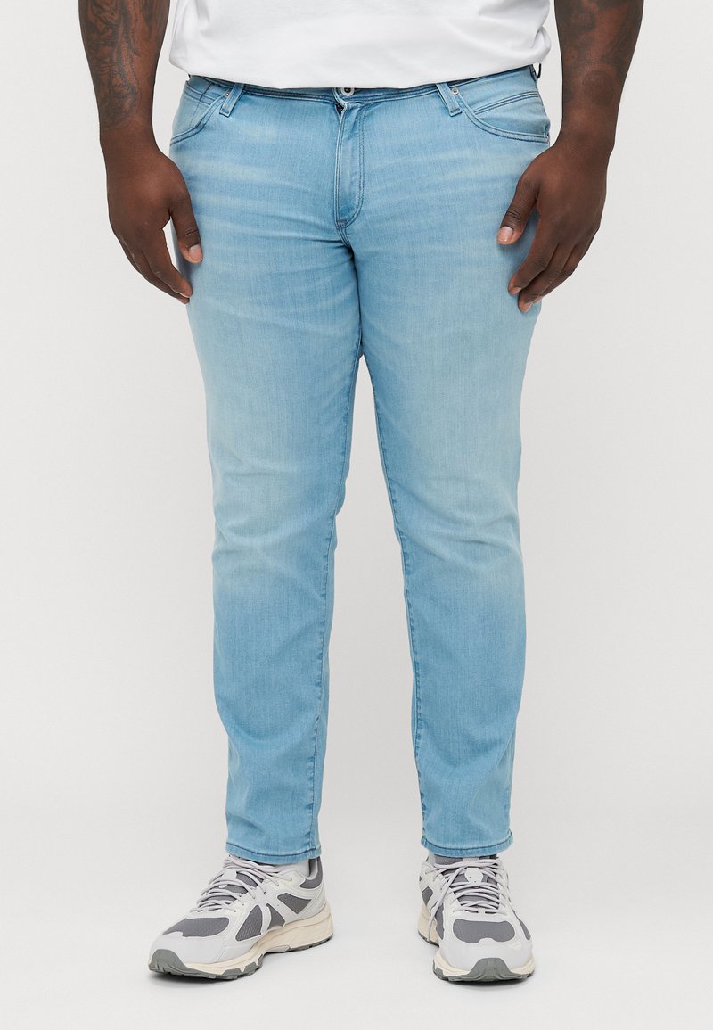 Jeans de mezclilla de color azul claro con un ajuste slim, con cremallera clásica, cinco bolsillos y una sutil textura deslavada. Combinados con zapatillas grises.