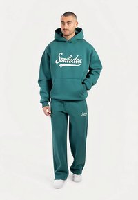 Sudadera con capucha verde azulado con el logo "Smilodox" en blanco y bolsillo tipo canguro, combinada con pantalones a juego. Hecha de material suave, cuenta con puños elásticos.