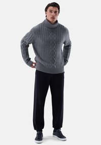 Grauer, gestrickter Rollkragenpullover mit texturiertem Rippenmuster und Zopfmuster, kombiniert mit dunklen marineblauen Hosen und grauen Sneakers.