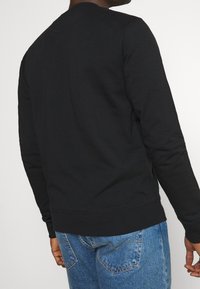 Felpa nera con maniche lunghe, polsini e orlo a costine, realizzata in tessuto morbido, indossata con jeans blu che mostrano cuciture dettagliate.