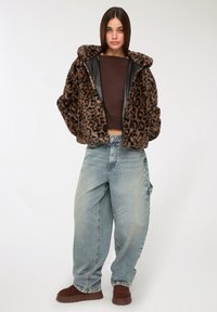Giovane donna che indossa una giacca in pelliccia finta con stampa leopardo, un top corto marrone, jeans larghi di un leggero blu, e scarpe slip-on marroni, in piedi con le mani nelle tasche.