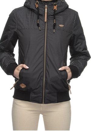 Frau in einer schwarzen Kapuzenjacke mit Zickzack- und Pünktchenmustern, die die Hände in den Taschen hat, kombiniert mit heller beiger Hose.