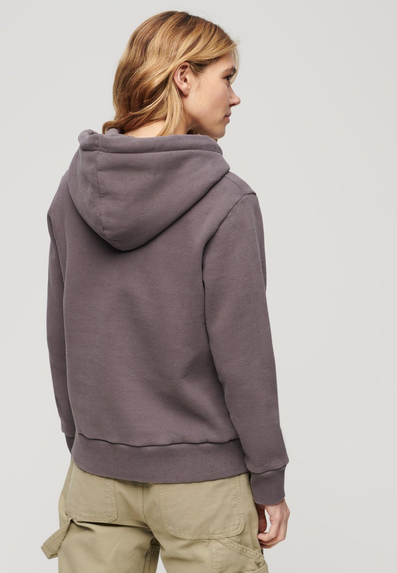 Kobieta z blond włosami do ramion, ubrana w fioletowy hoodie i beżowe spodnie typu cargo, odwrócona plecami na gładkim tle.