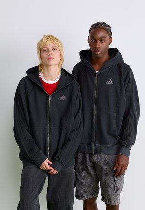 Dwaj mężczyźni noszą czarne rozpinane bluzy z kapturem w pasy, z logo Adidas. Jeden z nich ma pod spodem czerwoną koszulę.