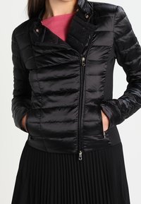 Femme portant une veste matelassée noire avec des poches zippées, superposée à un haut rose et assortie d'une jupe plissée noire.