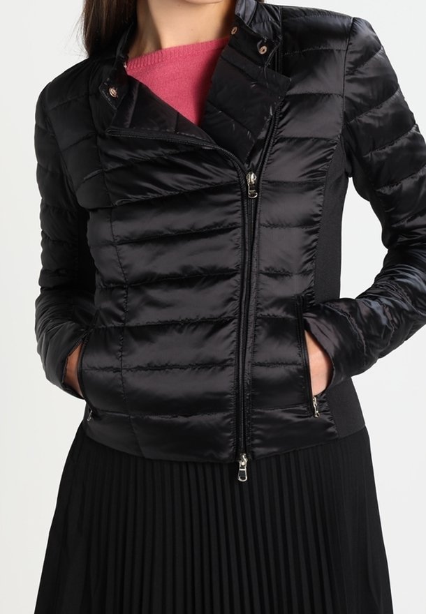 Femme portant une veste matelassée noire avec des poches zippées, superposée à un haut rose et assortie d'une jupe plissée noire.