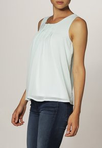 Top senza maniche verde chiaro con pieghe sul davanti, realizzato in tessuto morbido. L'orlo è leggermente curvato con una finitura liscia. Indossato con jeans scuri.