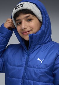 Modra podložena jakna z visokim ovratnikom, celim zadrgo in belim logotipom Puma. Model nosi svetlo sivo pleteno kapo. Gladka tekstura.