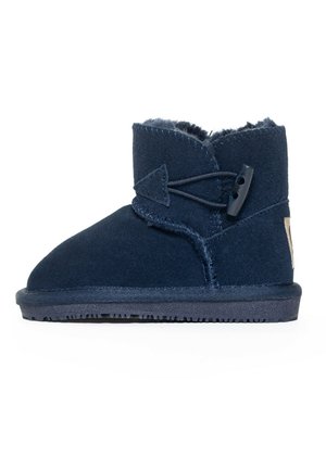 Bota azul marino con material suave, similar al fieltro, puntera redondeada y suela texturizada. Presenta una correa decorativa y cierre de botón.