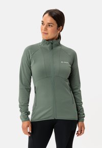 Veste ajustée verte avec un col montant, devant zippé et deux poches latérales. Tissu texturé avec un motif ribbé et une marque sur la poitrine.