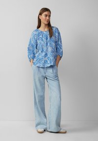 Blaue gemusterte Bluse mit rundem Ausschnitt und Dreiviertelärmeln, kombiniert mit hellblauen weiten Jeans und beigen Plateauschuhen.
